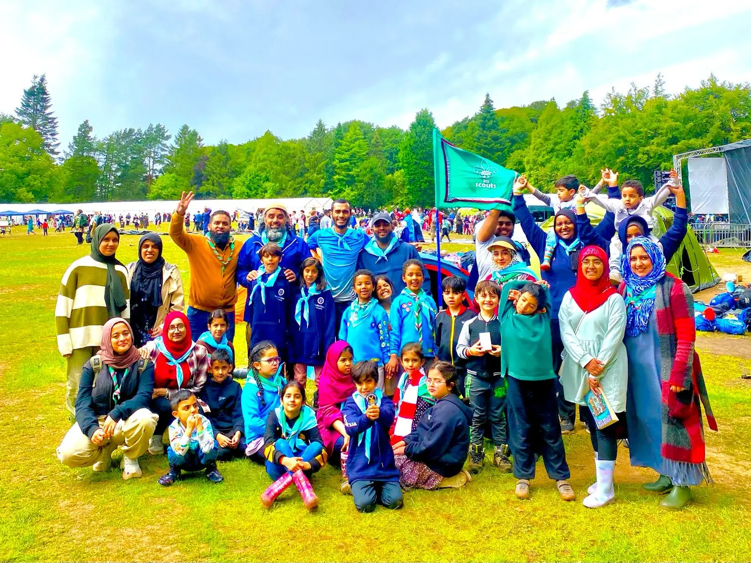 A diverse group of children and adult leaders in scout uniforms and casual outdoor clothing posing on a grassy field with tents and a forested background.