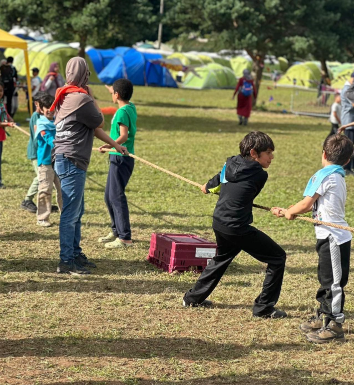 A group of children and an adult pulling on a rope in a grassy campsite, with colorful tents pitched in the background.
