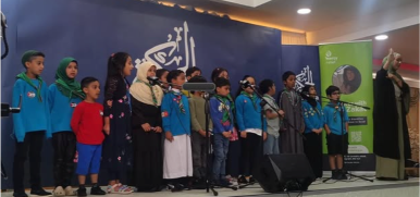 A diverse group of children and an adult woman stand in a row on a stage, many wearing bright blue scout hoodies with neckerchiefs and a few in traditional or casual attire.