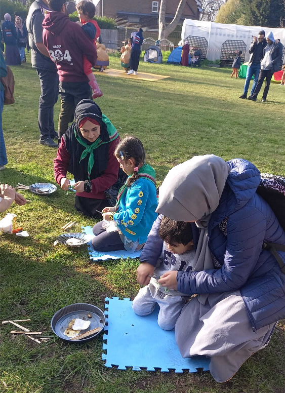 Two women wearing hijabs and two children seated on foam mats in a grassy park, arranging craft materials on paper plates; in the background, more families, tents, and community activities under a clear sky.