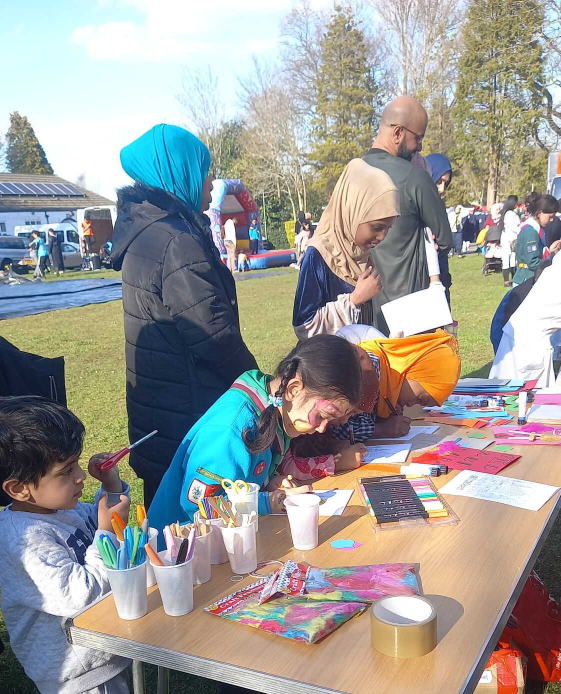 Children and adults gathered around a folding table in a sunny park, with cups of markers, paints, and colorful paper, one girl with a painted face leans over to create artwork while others including a toddler and adults in headscarves join in, and a bounce house and trees appear in the background.