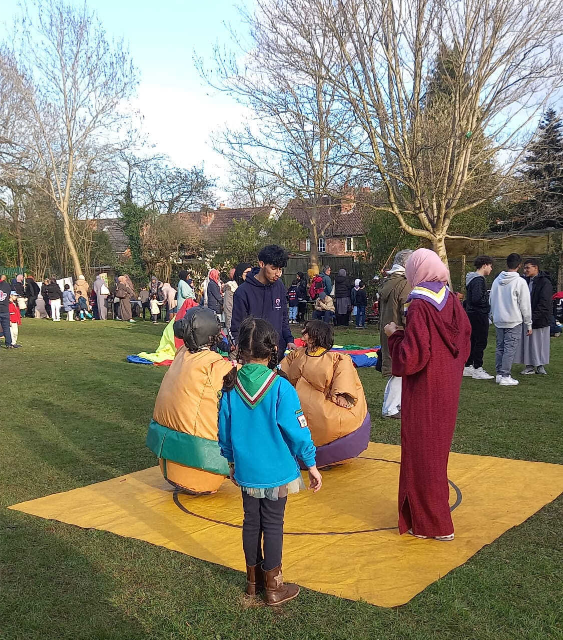 Children in inflatable sumo wrestling suits stand on a yellow mat in a grassy park, supervised by a volunteer in a maroon robe and hijab, while families and other participants mill about in the background.
