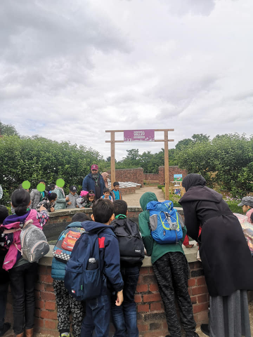 Children wearing colorful backpacks gathered around a low, circular brick feature beneath a wooden torii gate in a lush, overcast park setting, supervised by an adult.