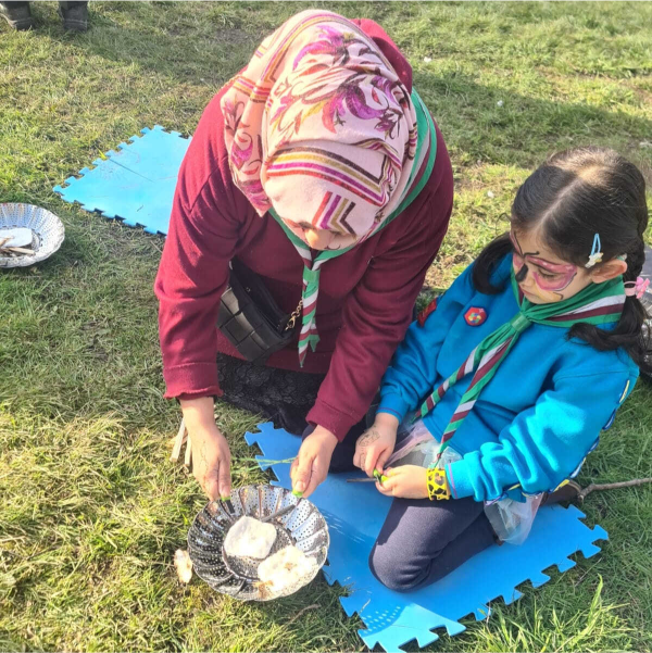 A scout leader wearing a patterned headscarf and maroon sweater kneels on a blue foam mat beside a young girl in a teal scout uniform and butterfly face paint. They hold metal strainer bowls over a small cooking setup on grass, shaping dough pieces together outdoors.