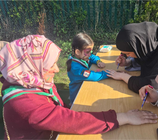A sunlit outdoor scene featuring three females around a wooden table in a park: a woman wearing a pink-patterned headscarf rests her hand on the table as another woman in a black hijab carefully paints her nails, while a young girl in a blue uniform with braided hair and face paint looks on.