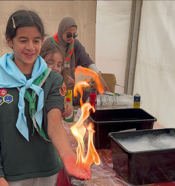 A girl in a scout uniform with a light blue neckerchief holds a small flame on her open palm inside a white tent, while two peers one wearing glasses and another in a headscarf observe from behind.