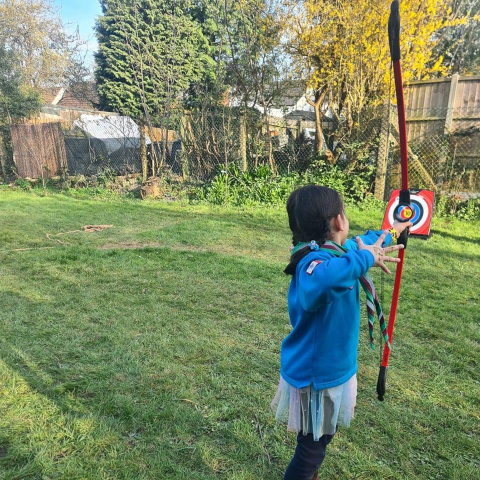 A young girl in a blue jacket and light skirt stands on a grassy lawn, drawing a red bow as she aims at a circular red and white target mounted on a stand, with trees and a wire fence in the background.
