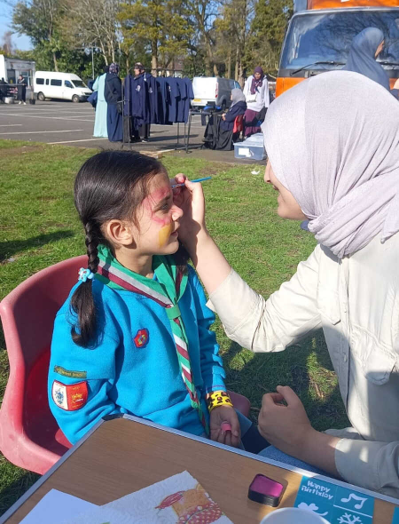 A young girl in a blue scout uniform sits on a red plastic chair outdoors while an adult woman wearing a light colored hijab uses a brush to apply pink and yellow face paint to her cheek; in the background, tables with painting supplies, other attendees, and parked vehicles underscore a lively community event.