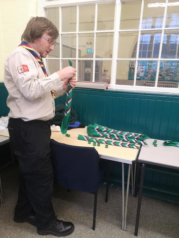 Adult scout leader in a beige uniform shirt and neckerchief inspects a green, white and red striped scarf at a table indoors, with more scarves laid out and tall windows in the background.