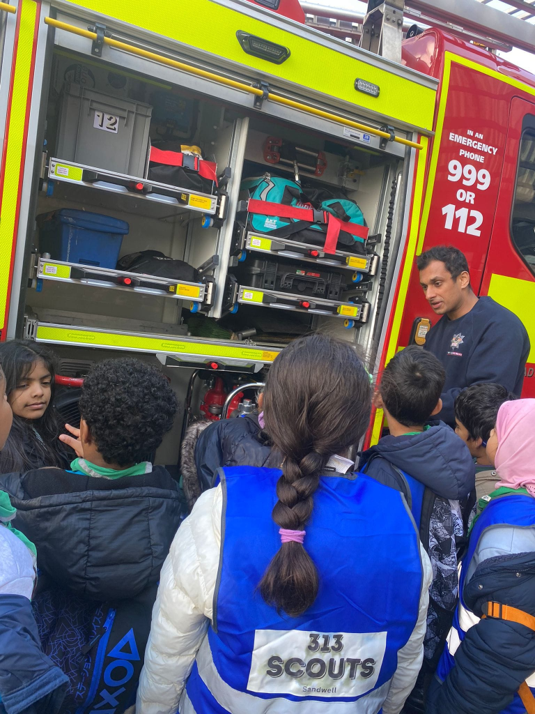 Children in blue “313 Scouts Sandwell” vests gathered around a firefighter showing the open compartments and equipment of a fire engine.