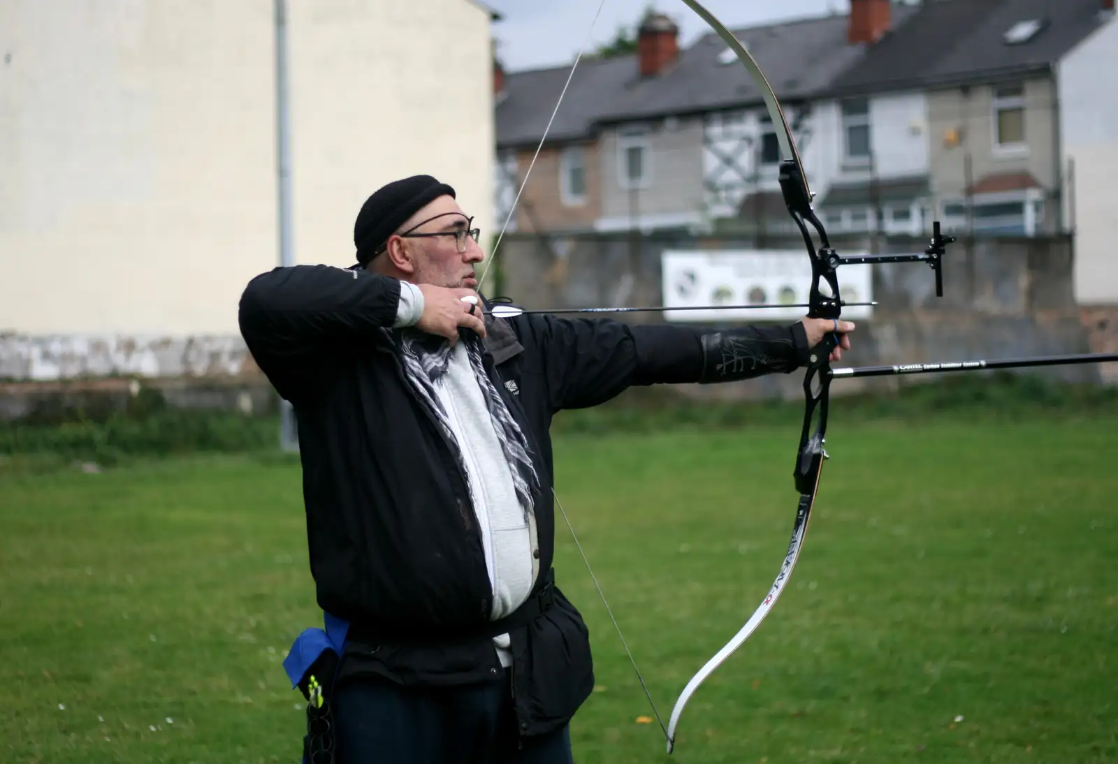 A man in a black jacket and beanie draws a recurve bow on a grassy field, aiming at an off-frame target.