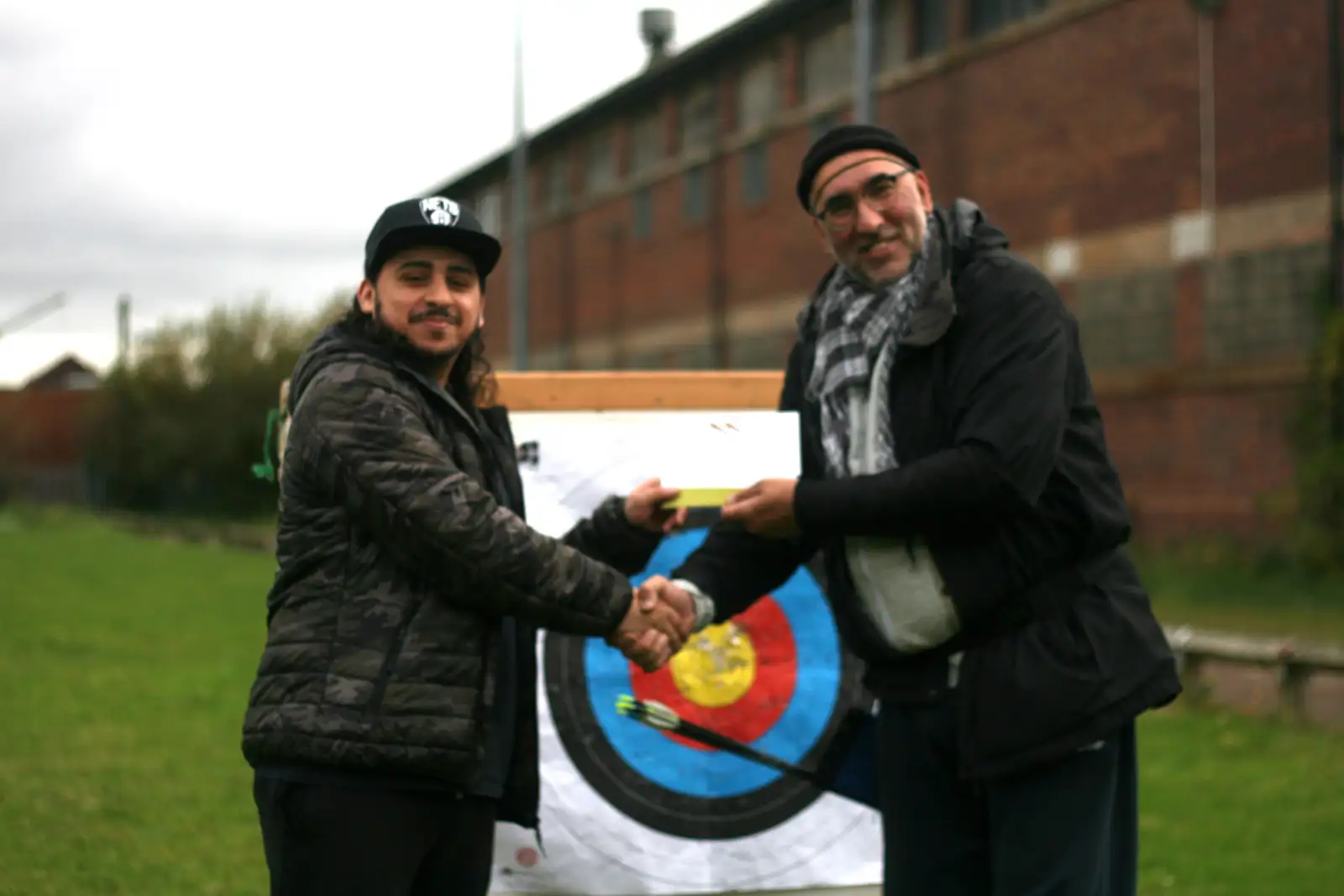 Two men shaking hands while holding an archery target with arrows embedded in it, standing outdoors in front of a brick building with green grass visible.