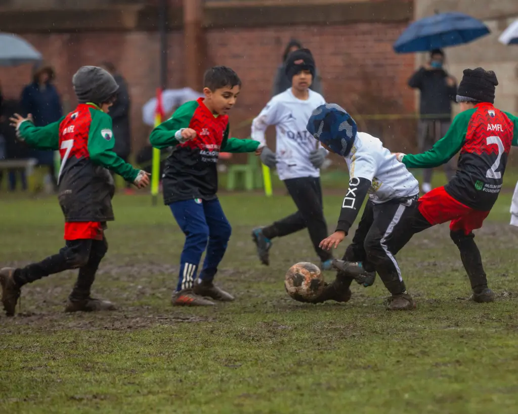 Children playing football match on a muddy field during rainy weather, showcasing their determination and enthusiasm.