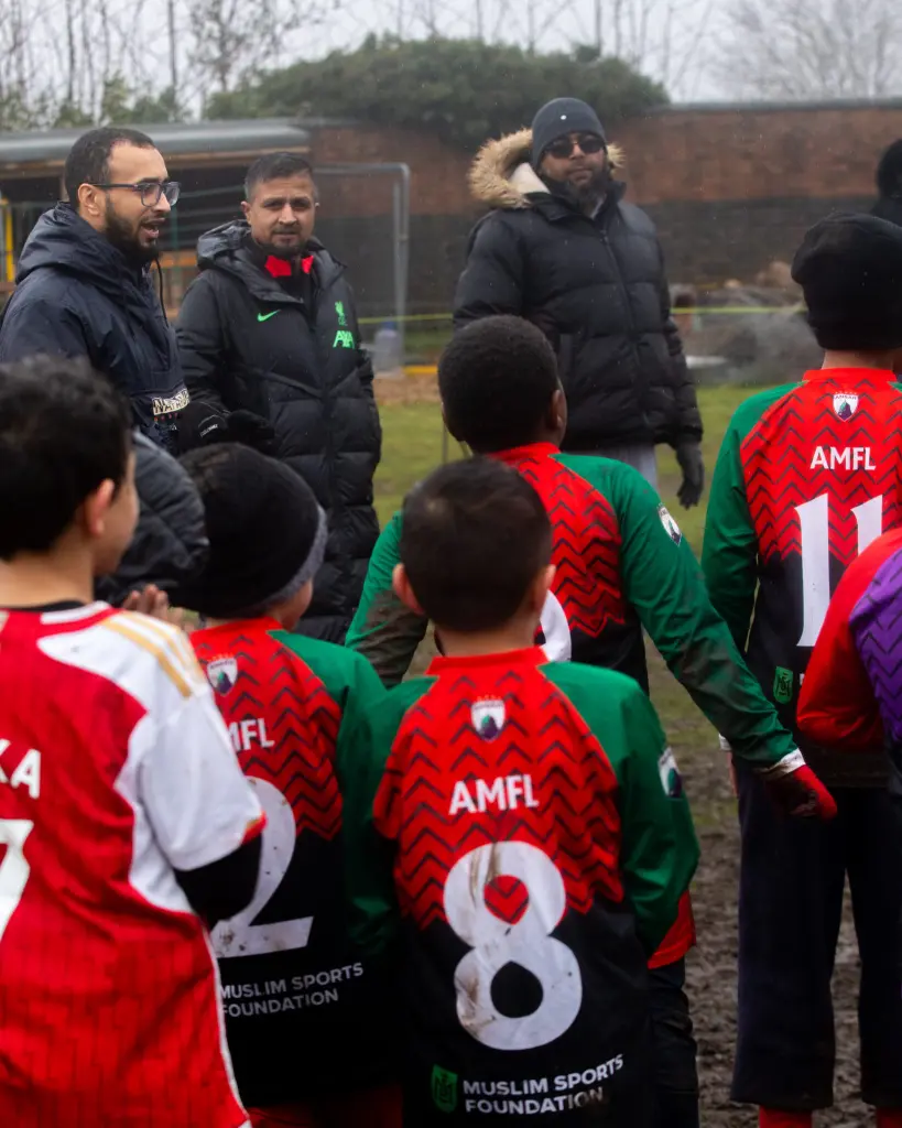 A group of young boys wearing red and green Muslim Sports Foundation football kits stand in a semi-circle on a muddy outdoor pitch, listening to three adult coaches dressed in winter jackets and hats on a cold, overcast day.