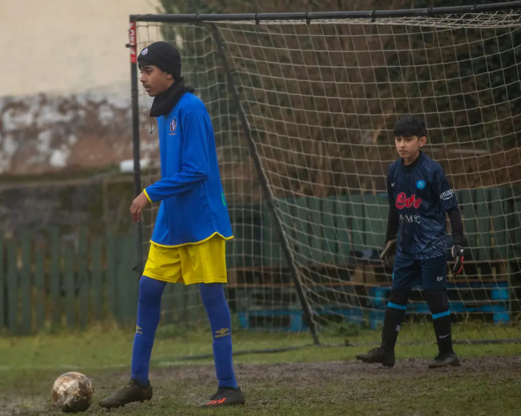 Two boys play football on a muddy outdoor pitch in cold weather. One boy in a blue long‑sleeve jersey, yellow shorts, blue socks, and a black hat is about to kick a dirty football near the goal, while a younger boy in dark goalkeeper kit and gloves stands alert in front of the net behind him.