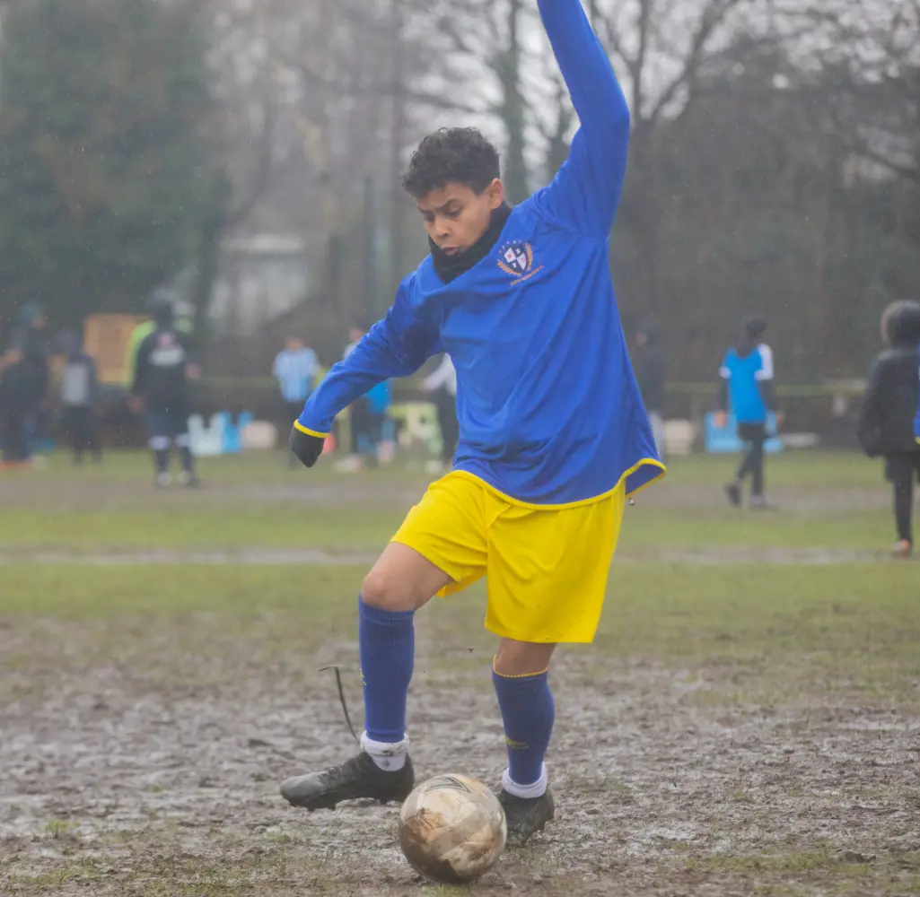 Football player in a blue jersey and yellow shorts swings his leg to kick a muddy ball on a rain soaked field, with blurred teammates and spectators in the background.