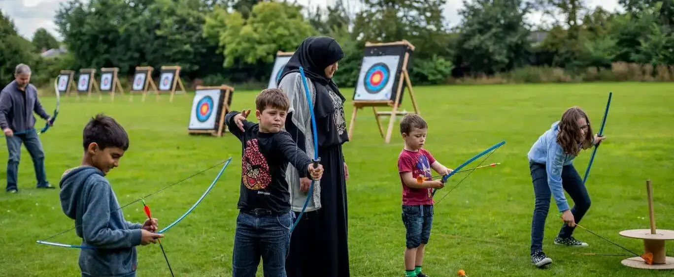 A group of children practicing archery under the supervision of an instructor in an outdoor setting, with targets visible in the background.