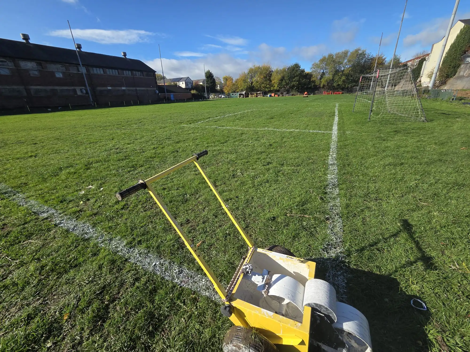A yellow and black line marking machine positioned on a well-maintained grass soccer field with white boundary lines, goal posts, and sports facility buildings in the background under a partly cloudy sky.