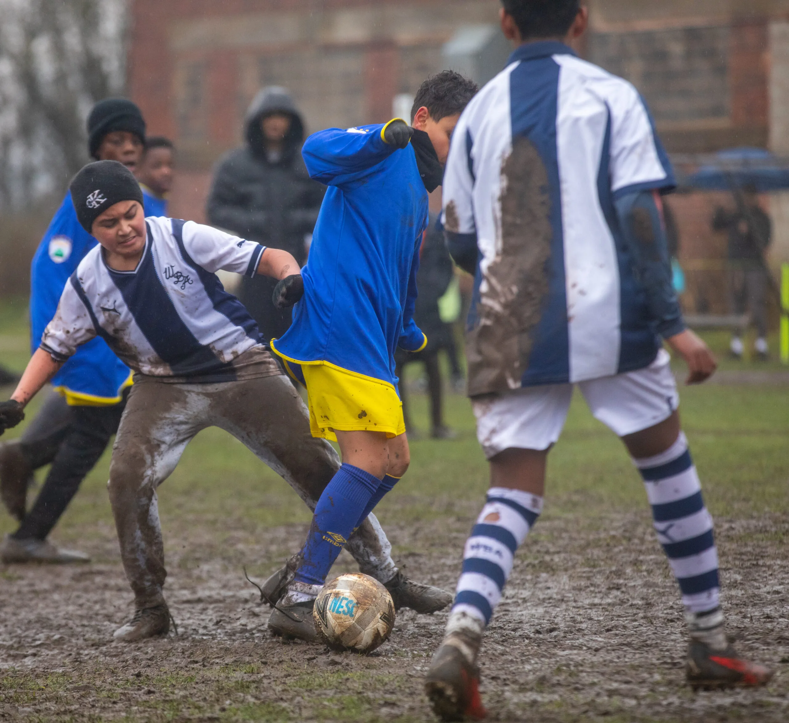 Three muddy youth soccer players contest the ball on a soaked field; a player in a blue shirt and yellow shorts shields the ball while two opponents in white-and-navy kits slide and challenge, with mud splattered over their legs and jerseys and blurred spectators in the rainy background.