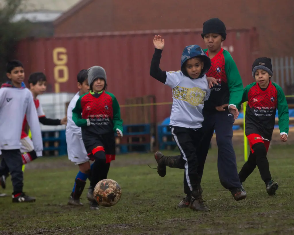 A group of young players, bundled in hats and jerseys, chase a muddy football across a wet, grassy field during a youth match.