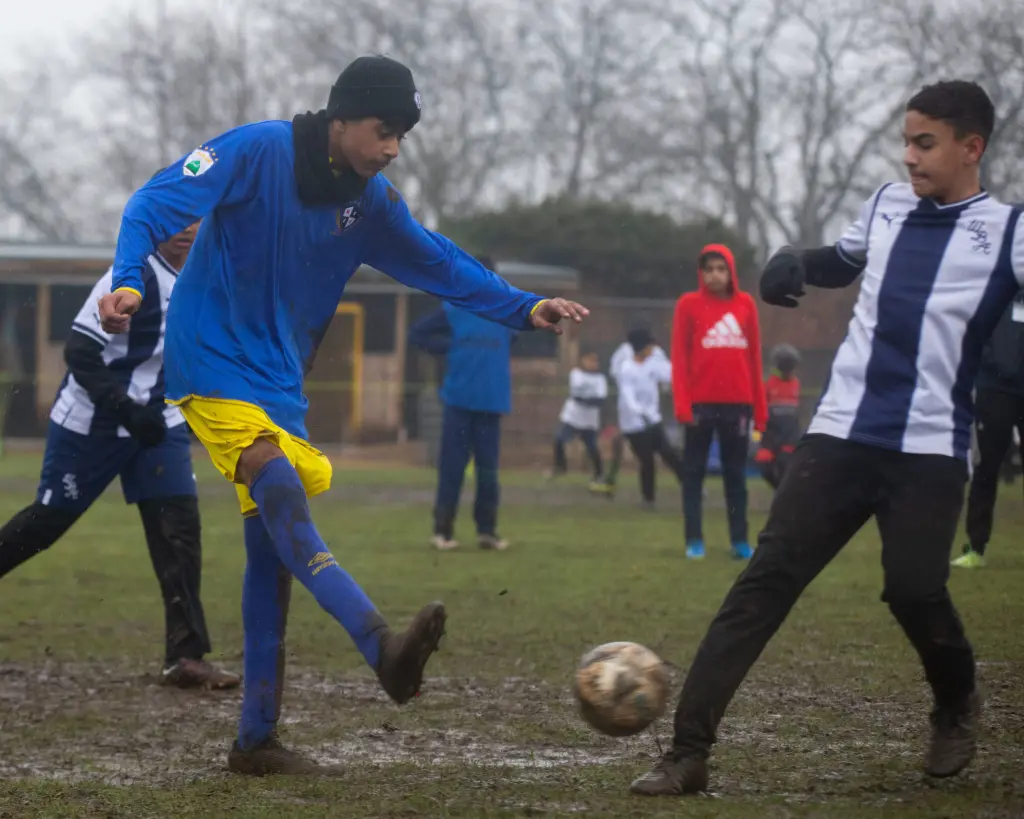 A player in blue and yellow strikes the ball while an opponent in white and blue challenges him on a wet, muddy pitch as teammates and spectators stand behind them.