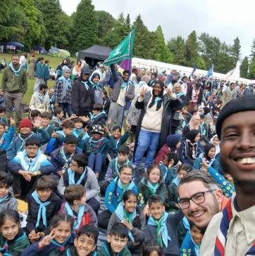 A large outdoor scout or community gathering with hundreds of children in blue uniforms and adults, gathered in an open field surrounded by trees with tents and colorful banners visible in the background.