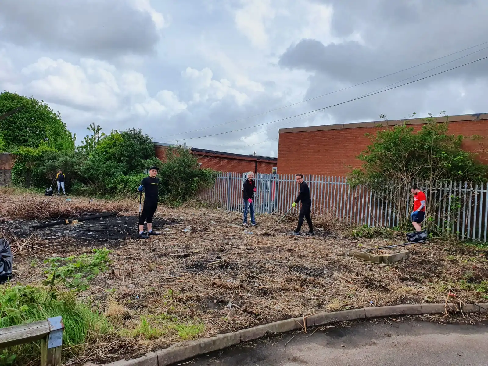Five volunteers in gloves using rakes and trash bags to clear dry vegetation and debris from a vacant lot bordered by a metal fence and brick building under a cloudy sky.