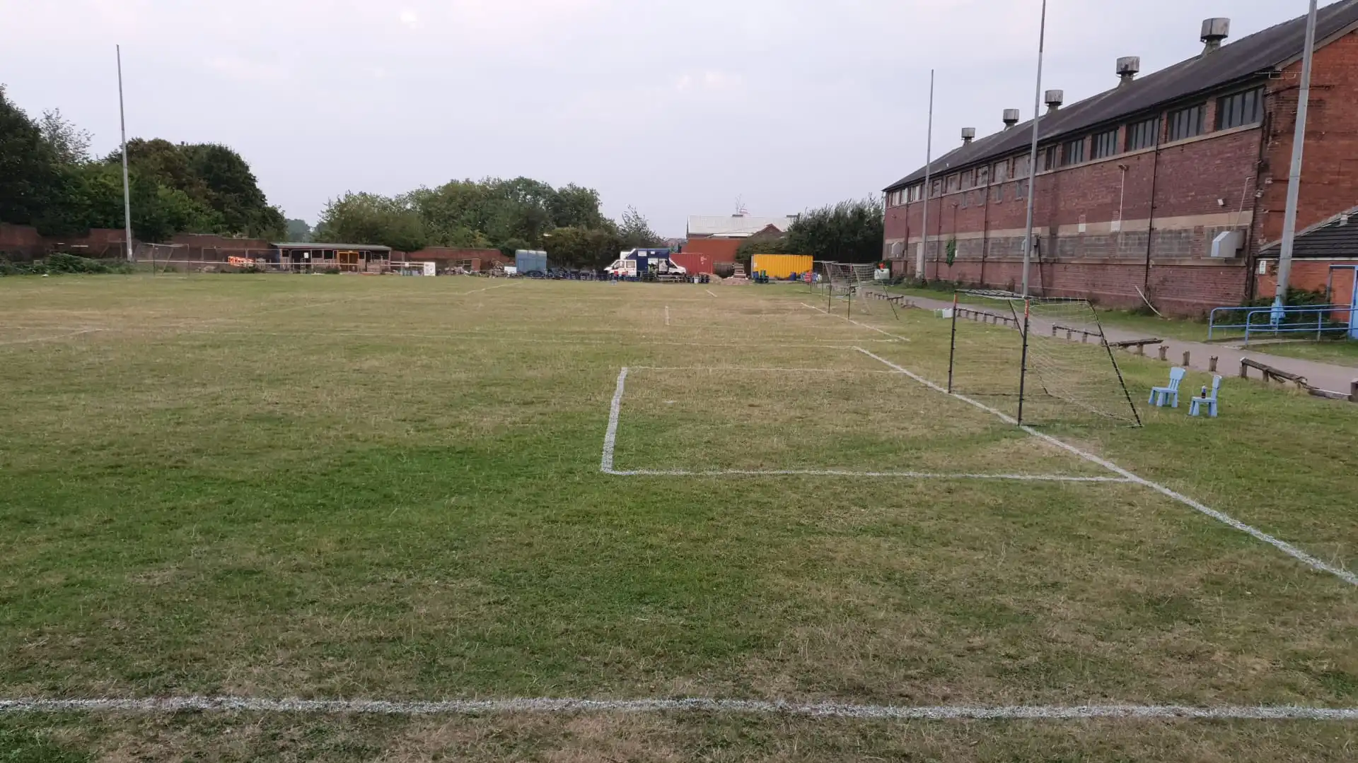 A community sports field with white boundary markings, hurdles, and blue chairs on the grass, bordered by a brick building with street lamps on the right, trailers and equipment storage on the left, and trees in the background under an overcast sky.