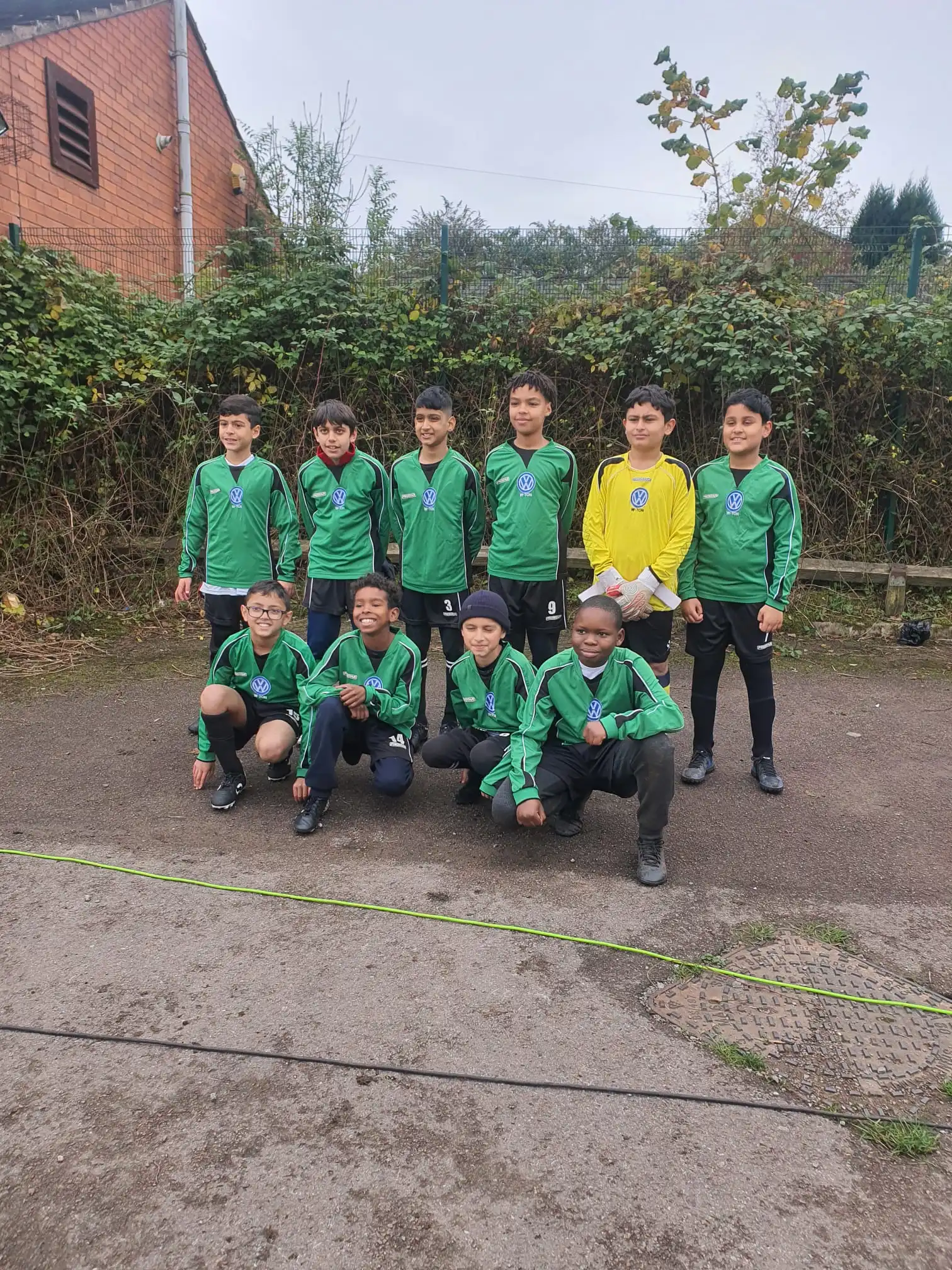 A youth soccer team of eleven boys wearing green jerseys (one goalkeeper in a yellow jersey) posing outdoors in two rows on a paved area in front of a leafy fence under an overcast sky.