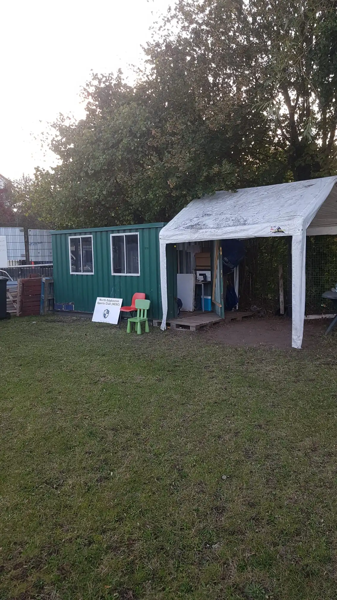 A grassy outdoor area with a green shipping container converted into a small office beside a white canopy tent, two small plastic chairs and a club sign sit in front, and trees form a backdrop.