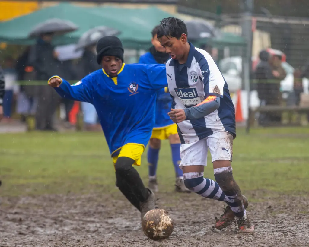 Two football players battle for the ball on a very muddy, rain soaked field. The player on the right wears a white and navy striped kit and is turning with the ball, their legs and uniform splattered with mud. The player on the left wears a blue jersey with yellow shorts and a black hat, stretching a leg toward the ball. Spectators holding umbrellas and a green canopy are visible in the blurred background.