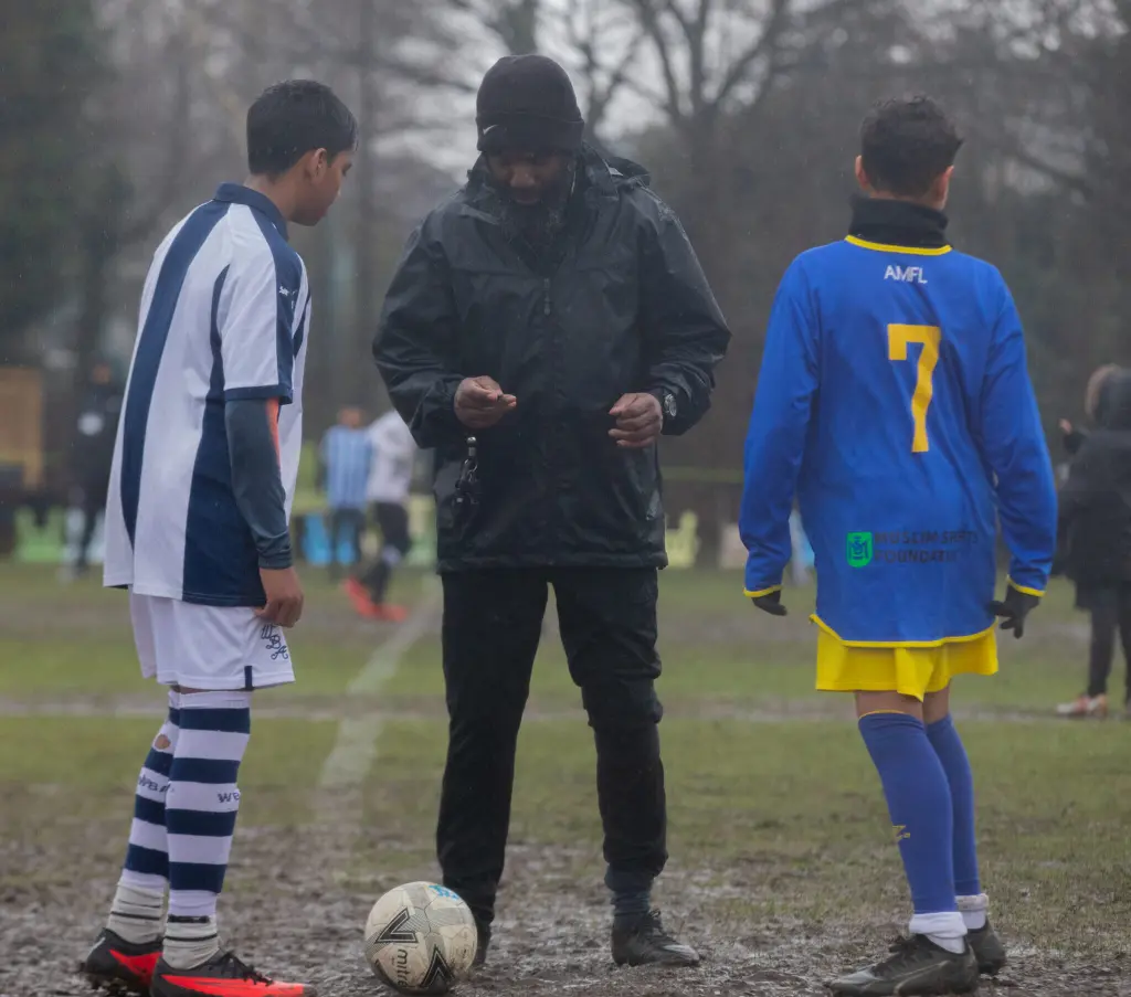 A referee in a black rain jacket and beanie flips a coin between two youth soccer players, one in white and navy stripes, the other in blue and yellow on a rainy, muddy pitch with a match ball at their feet.