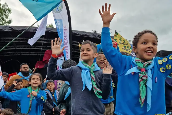 A group of children in blue scouts uniforms joyfully raising their hands at an outdoor event, with banners and flags in the background.