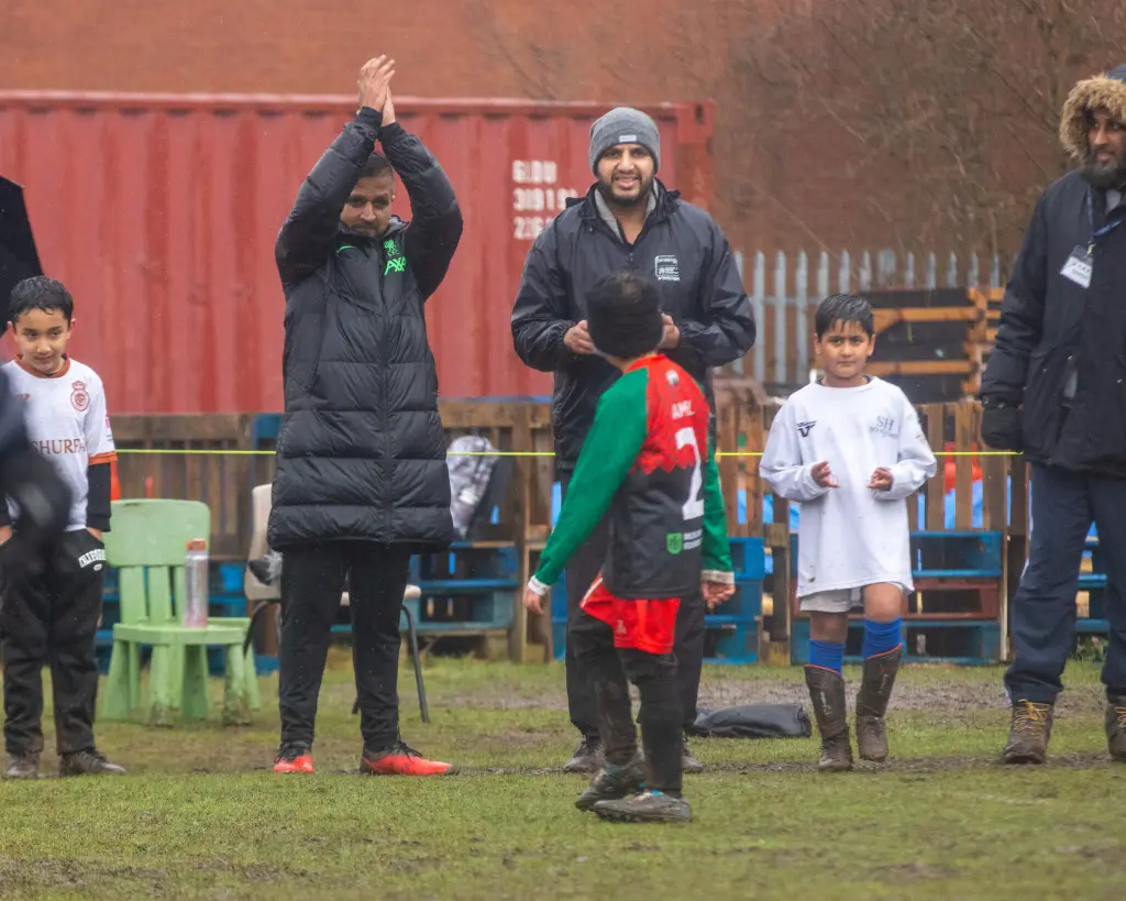 A muddy outdoor youth football scene: a boy in a red-and-green kit (number 2) stands front and center with his back to the camera while adults and other children on the sideline watch and applaud. Spectators wear rain jackets and boots; a red shipping container and wooden pallets form the background.