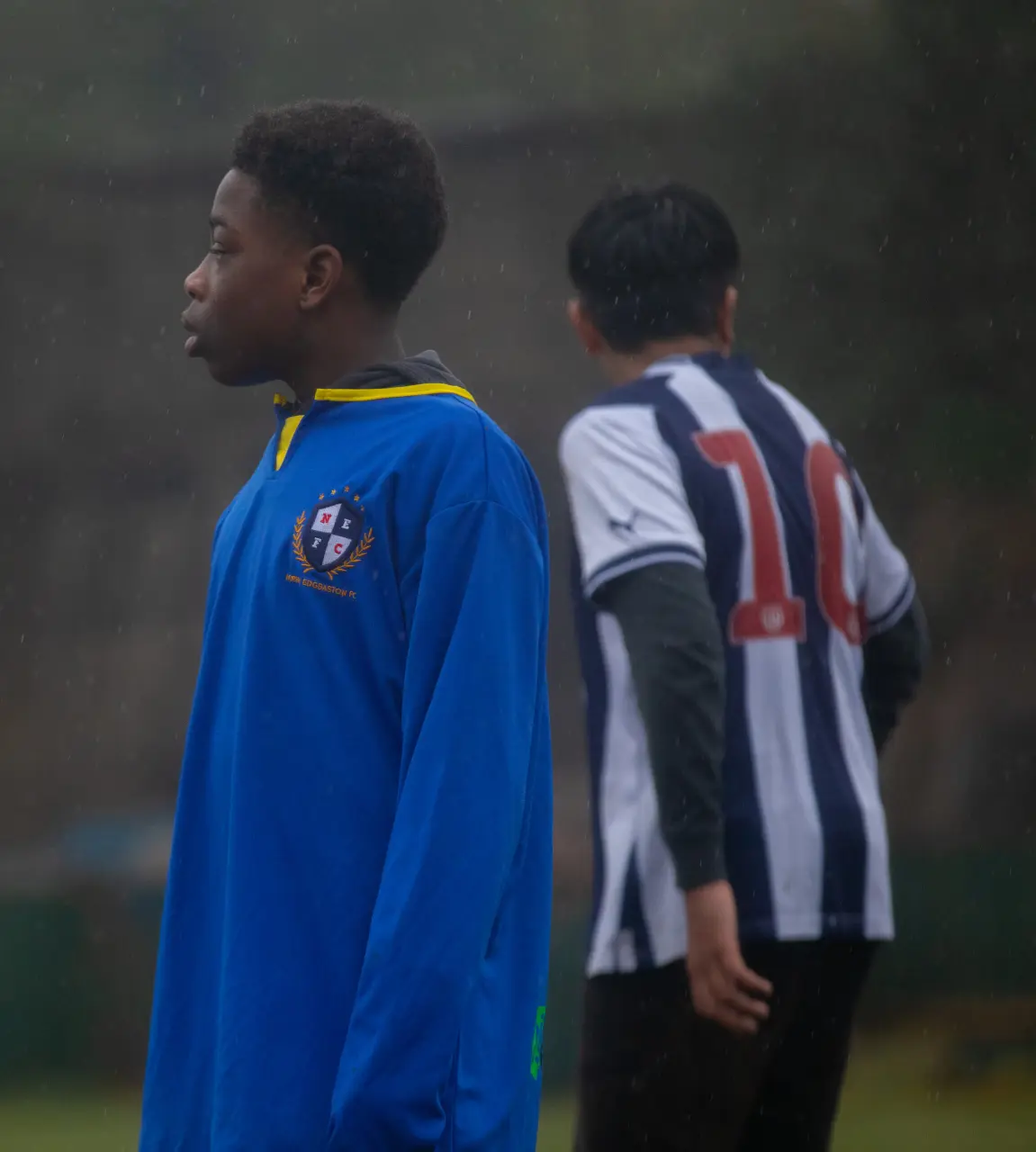 Two boys stand on a rainy outdoor sports field one in a blue long sleeve jersey with a crest, looking left, and the other in a black and white striped football shirt numbered 10, facing away.