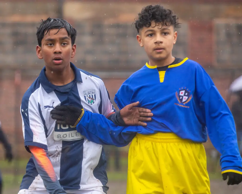 Two young boys compete for position in a rainy youth soccer match. The player on the left wears a navy-and-white West Bromwich Albion–style kit (with a Puma logo and mud stains), while the player on the right wears a blue jersey with yellow trim and yellow shorts, his hand resting on his opponent’s chest. Both look upward with focused expressions as raindrops fall, and the blurred background shows an overcast outdoor pitch.